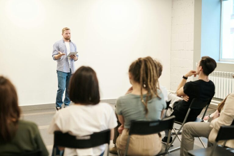 Man presenting with tablet to a small seated audience in a modern indoor setting.