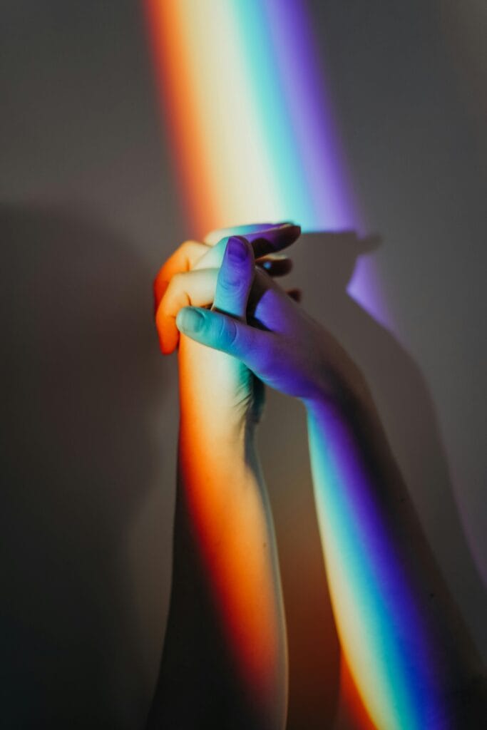 A pair of hands clasped together illuminated by a vibrant rainbow light, symbolizing LGBTQ+ pride and unity.
