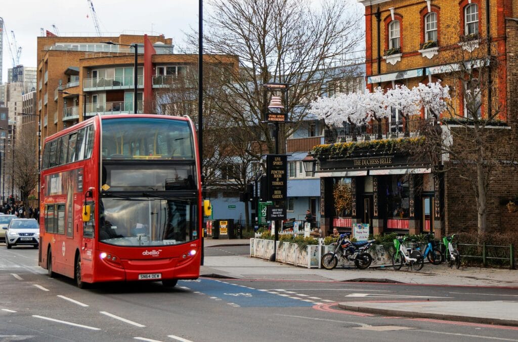 Iconic red double-decker bus on a bustling London street, passing by urban architecture.