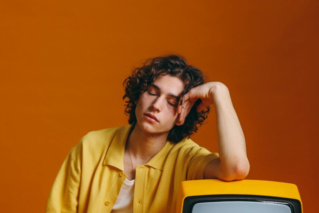 Young man with curly hair in vibrant yellow attire resting on a vintage TV set.