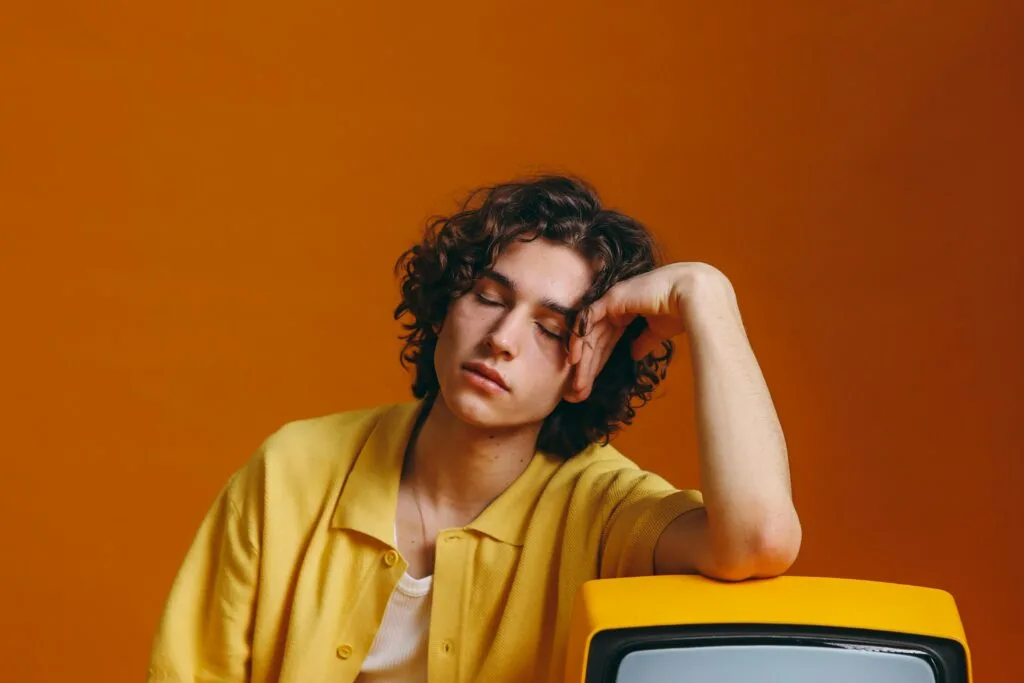 Young man with curly hair in vibrant yellow attire resting on a vintage TV set.